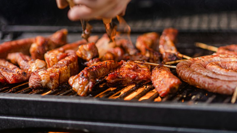 Vlees en worstjes op een rokende barbecue met open vuur, iemand keert het vlees met een tang