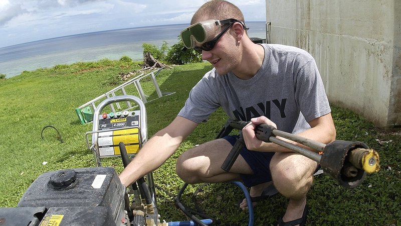 Man met lans en benzinemotor-hogedrukreiniger op een grasveld, klaar om een gevel schoon te spuiten